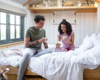 A young couple drinking coffee on a short queen mattress in their RV.